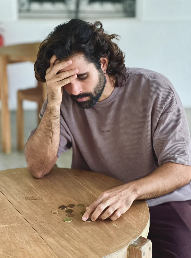 Homem sentado à mesa olhando para moedas, com a mão na testa